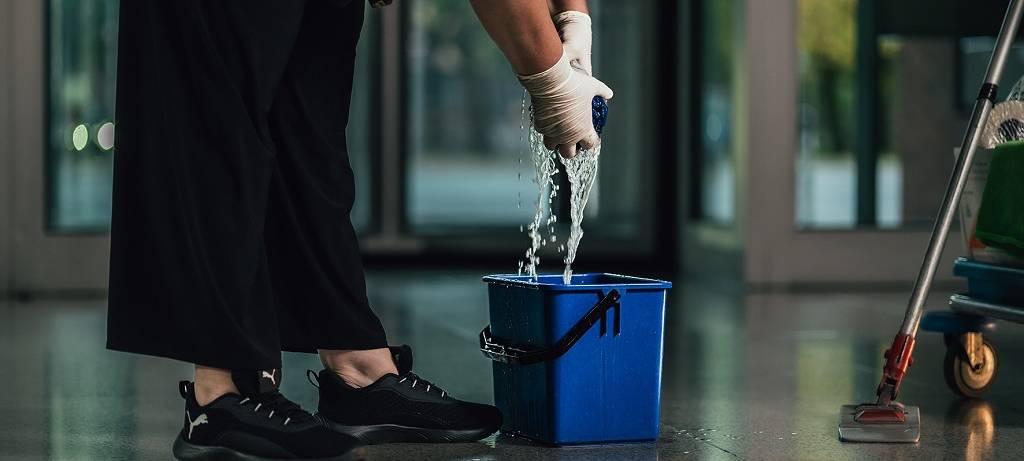 Detailaufnahme einer Gebäudereinigerin (Knie abwärts) in schwarzer Hose und schwarten Schuhen, die einen Wischlappen über einem Wischeimer auswringt. Rechts steht der Wischer angelehnt.