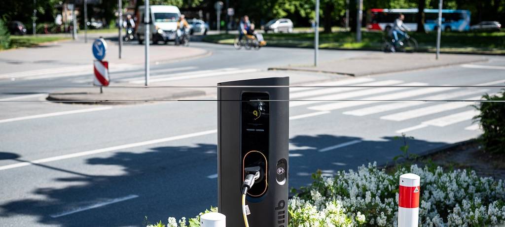 Bild einer Straßenkreuzung. Im Vordergrund eine E-Ladesäule mit eingestecktem Ladekabel. Davor die Front (Teilansicht) eines Autos.