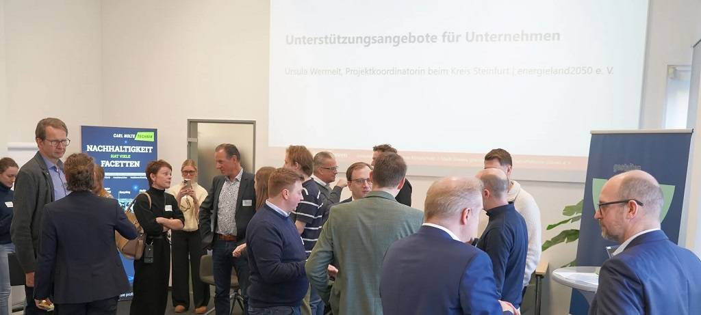 Teilnehmerinne und Teilnehmer der Unternehmensinitiative Klimaschutz im Gespräch. Im Hintergrund eine Leinwand-Präsentation und Aufsteller zum Thema.