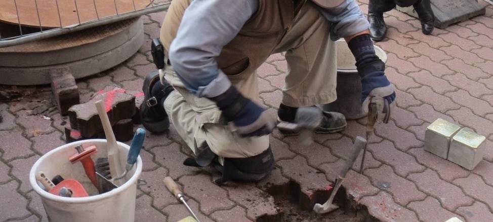 Mann mit Hut in Arbeitskleidung und Weste kniet vor aufgenommenem Straßenpflaster. Stolpersteine liegen bereit, um sie in das Loch einzusetzen.