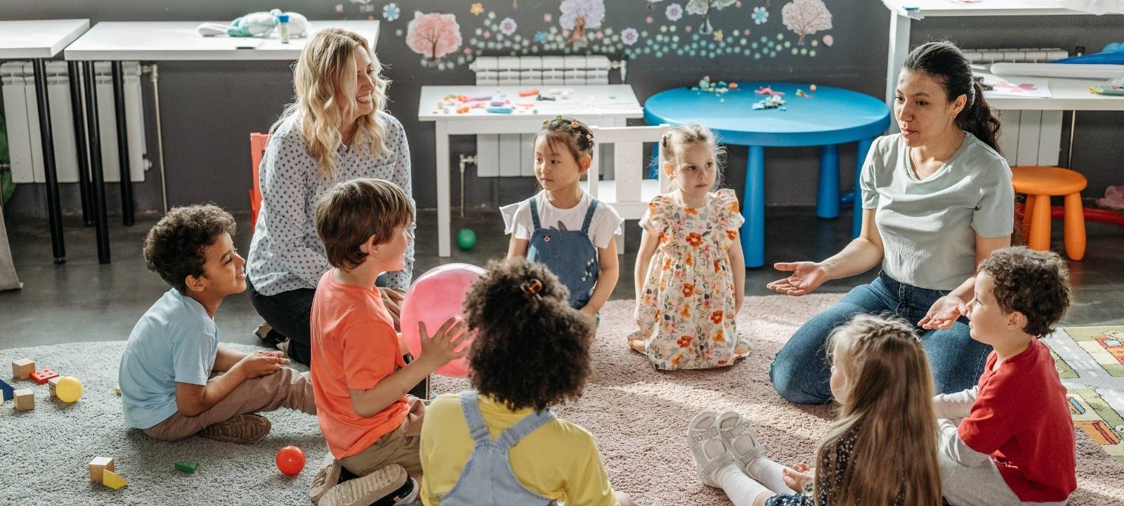 Erzieherinnen sitzen zusammen mit Kindern in einem Kreis auf dem Boden.