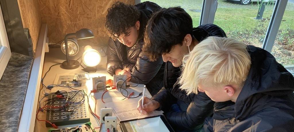 Drei Oberstufenschüler an einem Tisch voller Messegräte, Technik und Tablets. einer füllt eine Tabelle aus