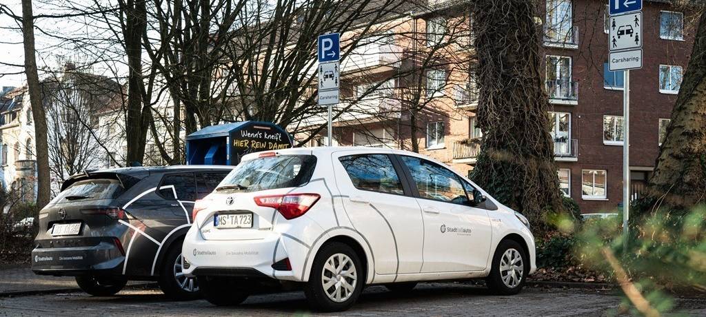 Weißes Carsharing-Auto auf einem Parkplatz vor einem Mehrfamilienhaus. Der Stellplatz ist durch Verkehrsschilder ausgewiesen.