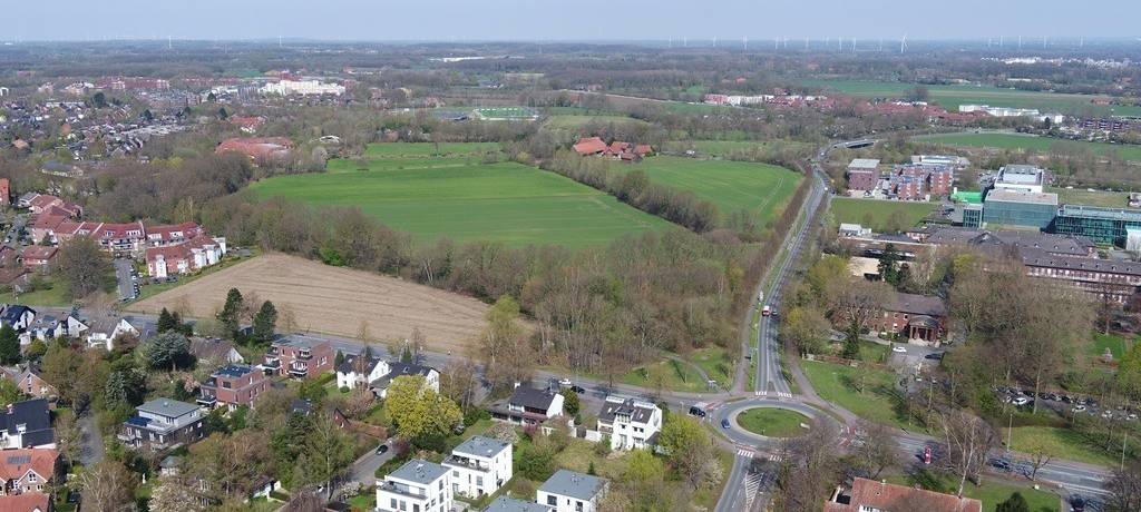 Luftbildaufnahme mit einer Freifläche, die von Bebauung fast umschlossen ist. Bis zum Horizont weitere lockere Bebauung und einige Windräder.
