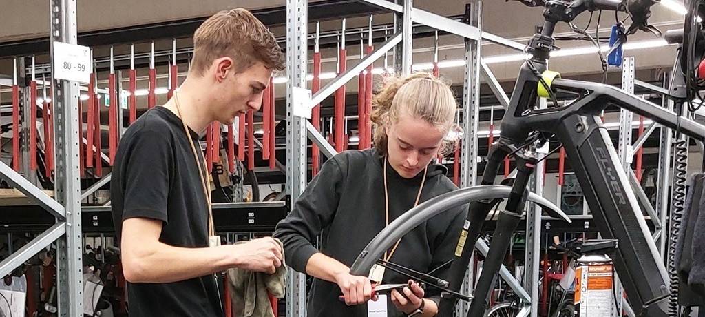 Junger Mann und junge Frau in Fahrradwerkstatt. Sie schraubt an einem Fahrrad, er steht dabei und erklärt.