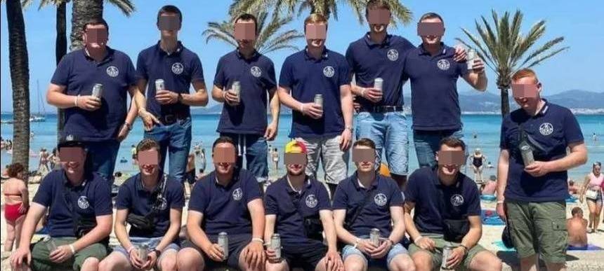 13 junge Männer in blauen T-Shirts auf einer Strandpromenade. Im Hintergrund Palmen, Wasser und Strand. Sechs sitzen im Vordergrund auf einer Betonabgrenzung, die anderen stehen. Alle haben eine Bierdose in der Hand. Die Gesichter sind verpixelt.