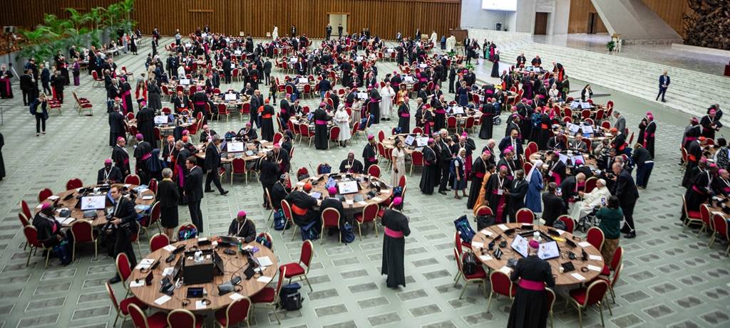 Blick von oben auf die Weltsynode 2024 in Rom. Ein großer Raum voller runder Tische, an denen Bischöfe und andere Kirchenfunktionäre konferieren.