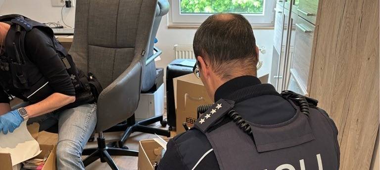 Zwei Zollbeamte in Uniform und mit blauen Nitrilhandschuhen durchsuchen einen Raum mit Kartons und Schreibtisch