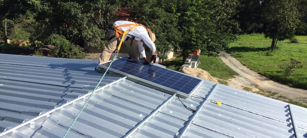 Ein Mann montiert ein Solarpaneel auf einem Dach, das mit Trapezblechen eingedeckt ist, vermutlich ein Hallendach. Er trägt ein langarmiges weißes Hemd und hat einen Strohhut gegen die Sonne auf. Im Hintergrund Bäume auf einer grünen Wiese, über die ein Weg führt.