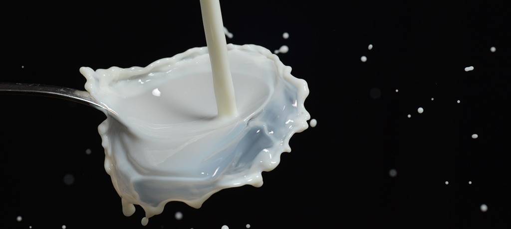 Ein Milchstrahl, der vor schwarzem Hintergrund auf einen Löffel trifft. Die Milch spritzt und tropft in alle Richtungen.