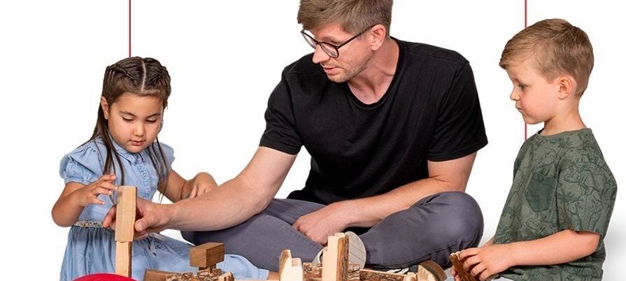 Junger Mann mit zwei Kleinkindern (Mädchen und Junge) beim Bauen mit selbstgemachten Holzbauklötzen.