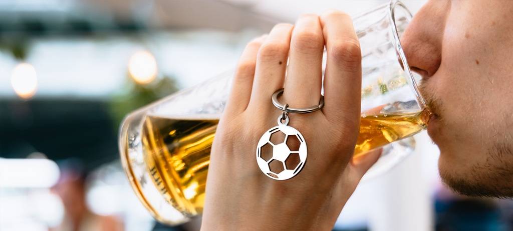 Close-Up auf jungen Mann, der Bier aus einem Glashumpen trinkt. Am Finger hängt ein Blech-Schlüsselanhänger mit Fußball. Ums Handgelenk trägt er ein Flechtarmband in Deutschlandfarben. Vom Gesicht sind nur Kinnpartie, Mund und Nase zu sehen.