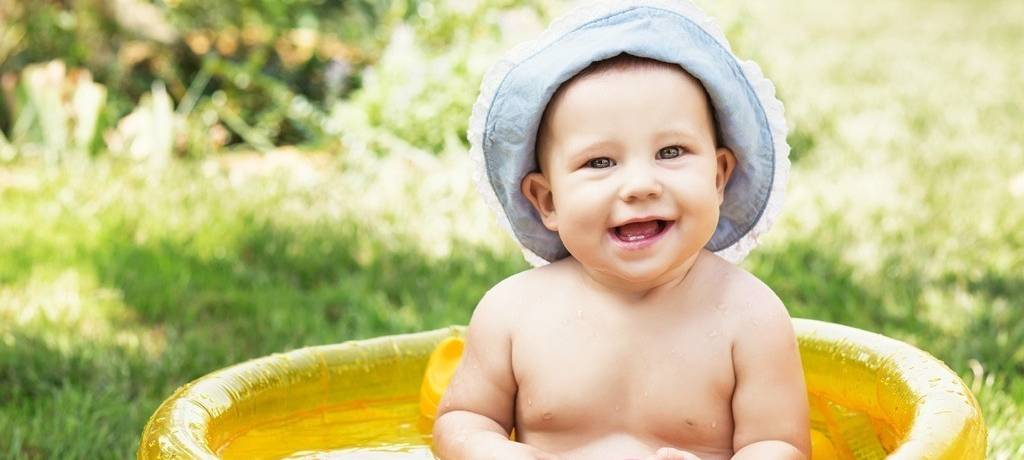 Baby sitzt in kleinem gelben Planschbecken auf dem Rasen, hat eine Mütze auf, ein Spielzeug in den Händen und lacht.
