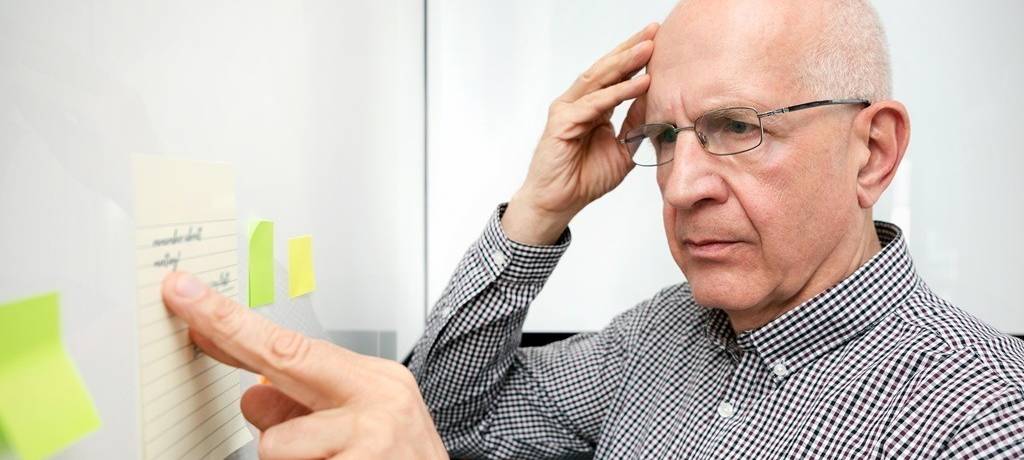 Älterer Mann mit Brille, Glatze und Karo-Hemd vor einer Pinnwand mit Notiz-Zetteln und Post-Its greift sich mit ratlosem Blick an den Kopf.
