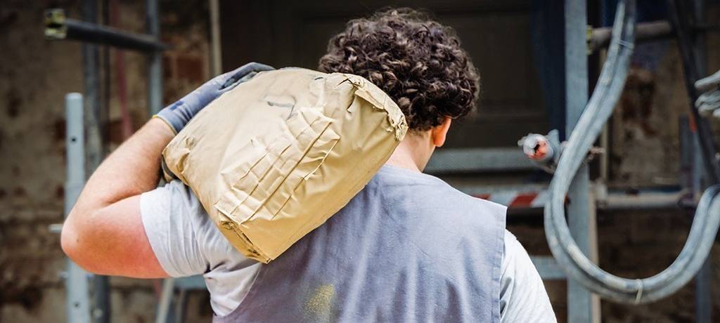 Junger Mann mit dunklem Lockenkopf trägt einen Sack (Mörtel?) auf der Schulter. Er ist von hinten zu sehen und trägt eine hellgraue Arbeitsweste und ein weißes T-Shirt.