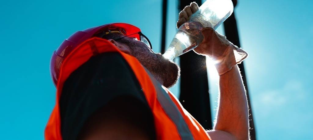 Arbeiter in orangener Arbeitskleidung und mit Bauhelm vor einem Kran trinkt aus einer Wasserflasche. Das Bild ist von unten aufgenommen, die Sonne leuchtet in der Flasche.
