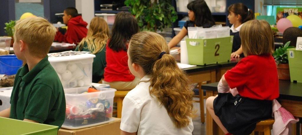 Schulkinder in einer alternativ wirkenden Lernsituation mit schuluntypischen Tischen, Stühlen und Lernmaterialien. Alle schauen auf etwas, das sich außerhalb des Bildes befindet.