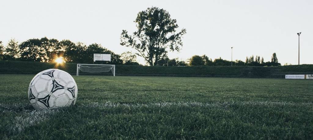 Abendlicher Sportplatz mit grünem Rasen, Fußball im Vordergrund und Baum und Sträuchern im Hintergrund vor bedecktem Himmel