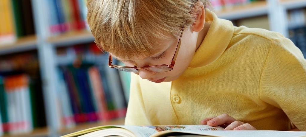 Kleiner Junge im gelben Polo und mit Brille schaut konzentriert und mit wenig Abstand in ein Buch. Im Hintergrund ein Bücherregal mit bunten Buchrücken.