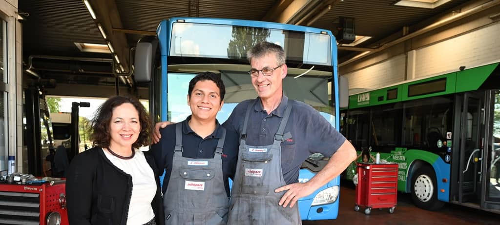 Blick in eine Bus-Werkstatt mit Bussen, Grube und Werkzeugwagen. Im Vordergrund zwei Männer in Arbeitskleidung mit Namensschilder und Aufschrift Schäpers Arm untergehakt mit einer Frau in T-Shirt, Rock und Jacke. Alle lächeln.