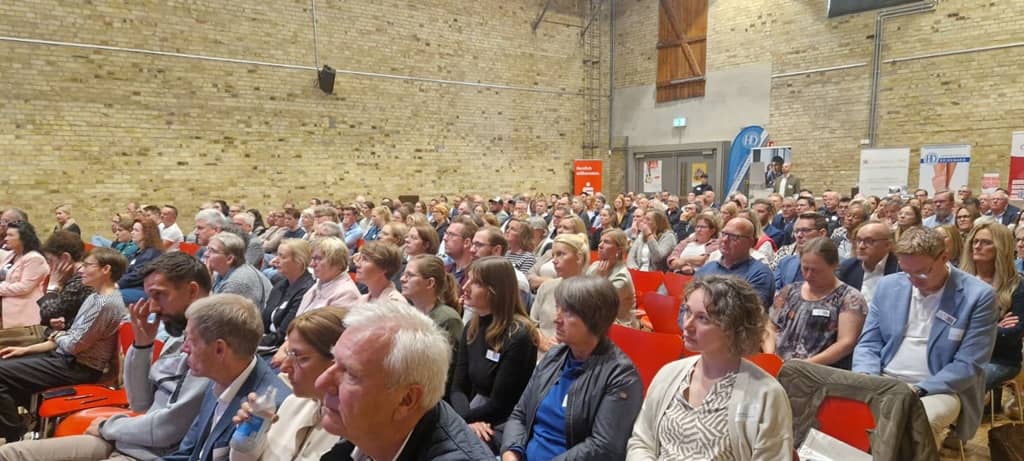 Teilnehmer einer Konferenz in Stuhlreihen in einem Saal blicken in eine Richtung (vermutl. zur Bühne).