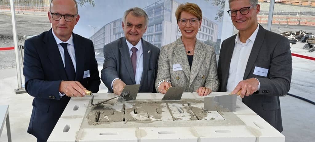 Drei Männer und eine Frau vor einem frisch gemauerten Block aus Kalksandsteinen mit Maurerkellen in den Händen zelebrieren die Grundsteinlegung, indem sie Mörtel in Fugen streichen.
