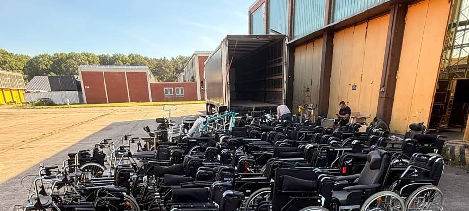 Ein Platz vor einer Halle, daruf ein LKW mit offener Ladeklappe. Davor eine große Anzahl von Rollstühlen.