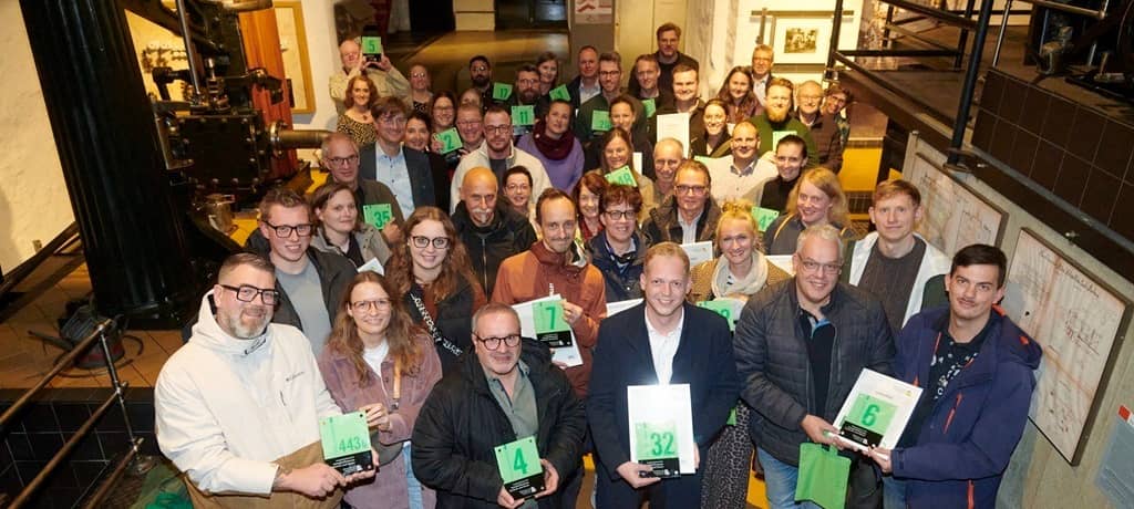 29 Männer und Frauen in einem Raum des Museums für Industriekultur halten lächelnd Tafeln mit grünen Hausnummern auf hellgrünem Hintergrund in den Händen