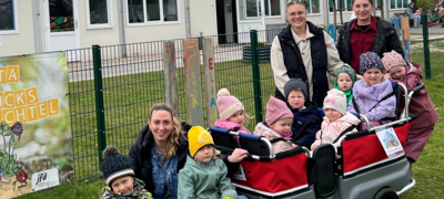Kindergartenkinder und Betreuerinnen mit Bobby Car, Dreirad und Bollerwagen auf einer Rasenfläche vor ihrer Kita